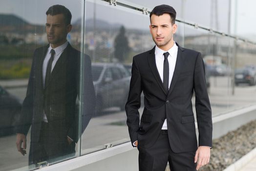 Portrait of a young businessman near a office building wearing black suit and tie with modern haircut