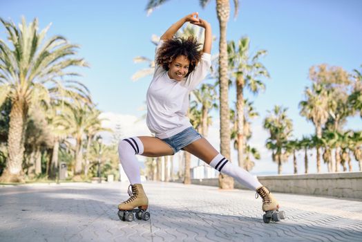 Smiling black woman on roller skates riding outdoors on beach promenade with palm trees. Smiling girl with afro hairstyle rollerblading on sunny day.