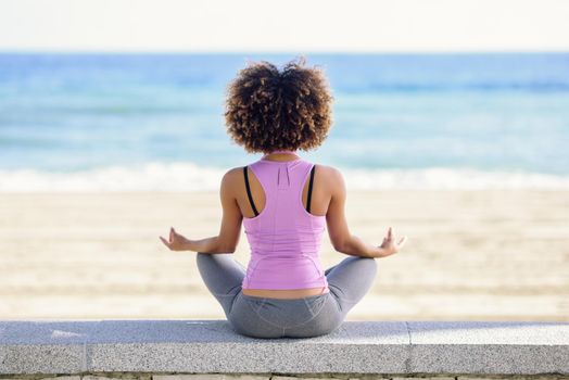 Rear view of black woman, afro hairstyle, doing yoga in the beach. Young Female wearing sport clothes in lotus figure with defocused background.