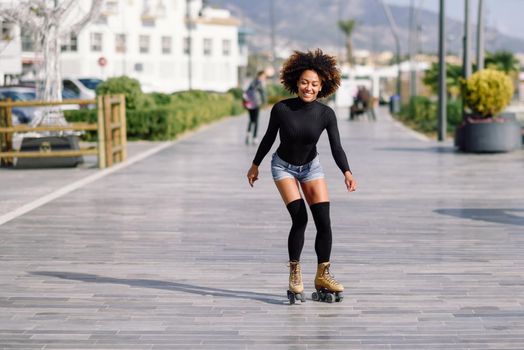 Young fit black woman on roller skates riding outdoors on urban street. Smiling girl with afro hairstyle rollerblading on sunny day