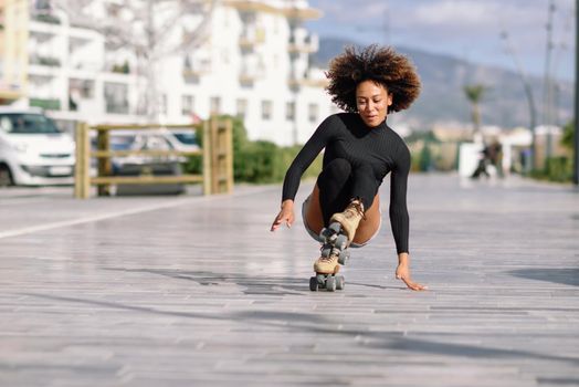 Young fit black woman on roller skates riding outdoors on urban street. Smiling girl with afro hairstyle falling to the ground.