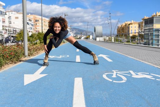 Young fit black woman on roller skates riding outdoors on bike line. Smiling girl with afro hairstyle rollerblading on sunny day