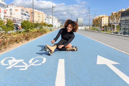 Young smiling black girl sitting on bike line and puts on skates. Woman with afro hairstyle rollerblading on sunny day