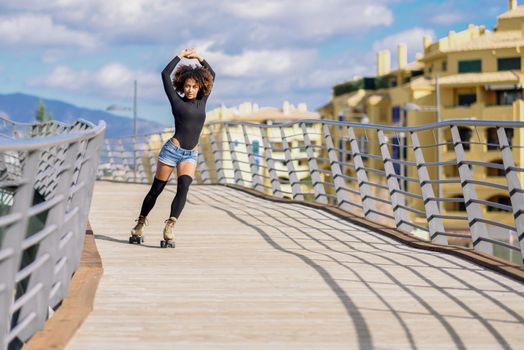 Young fit black woman on roller skates riding outdoors on urban bridge with open arms. Smiling girl with afro hairstyle rollerblading on sunny day. Beautiful clouds in the sky.