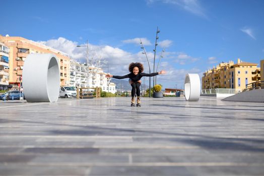 Young fit black woman on roller skates riding outdoors on urban street. Smiling girl with afro hairstyle rollerblading on sunny day