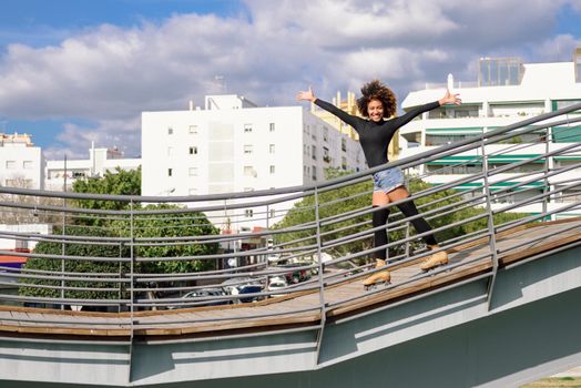 Young fit black woman on roller skates riding outdoors on urban bridge with open arms. Smiling girl with afro hairstyle rollerblading on sunny day. Beautiful clouds in the sky.