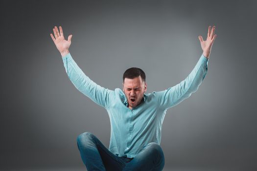 Casual young man sitting on the floor with his legs crossed and cheering with his hands in the air while screaming. Isolated on gray background