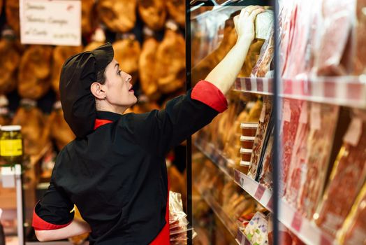 Portrait of female worker taking products in butcher shop. Refrigerated display case for sausage packages, ham and cheese