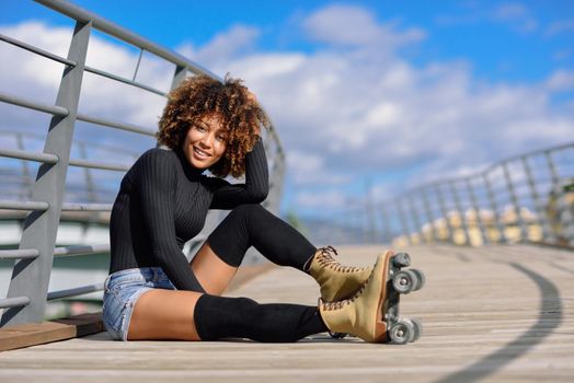 Black woman, afro hairstyle, on roller skates sitting outdoors on urban bridge. Smiling young female with beautiful clouds at the background.