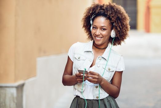 Young attractive black woman in urban street listening to the music with headphones and smartphone. Girl wearing casual clothes with afro hairstyle