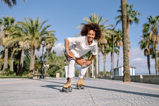 Smiling black woman on roller skates riding outdoors on beach promenade with palm trees. Smiling girl with afro hairstyle rollerblading on sunny day.