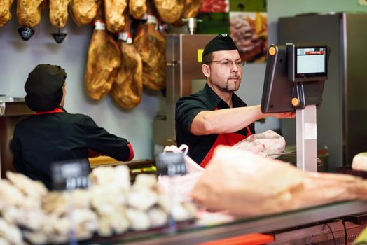 Butcher in a butcher's shop weighing the meat and charging with ham at the background