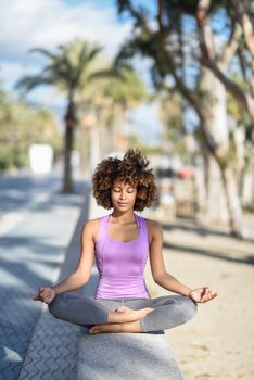 Black woman, afro hairstyle, doing yoga asana in the beach with eyes closed. Young Female wearing sport clothes in lotus pose with defocused background.