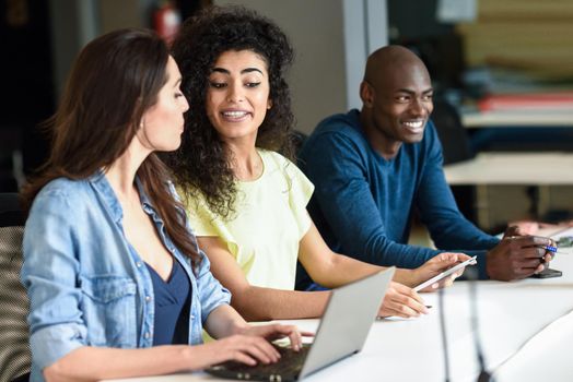 Three young people studying with laptop computer on white desk. Beautiful girls and man working toghether wearing casual clothes. Multi-ethnic group.