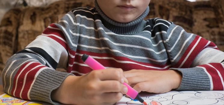 A little boy paints a coloring with crayons and felt-tip pens on a wooden table at home.