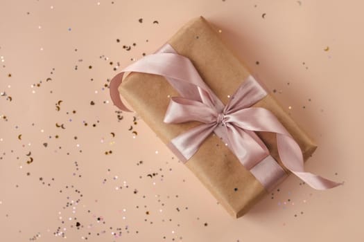 Birthday or New Year's eve present in kraft paper with pink ribbon on a soft beige background with glitter and confetti. Xmas composition. Flat lay. Happy holidays celebration and giving love concept