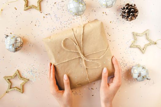 Female hands with natural manicure holding present in kraft paper with jute on soft beige background. Xmas composition. Flat lay. Happy holidays, New Year celebration and giving love concept.