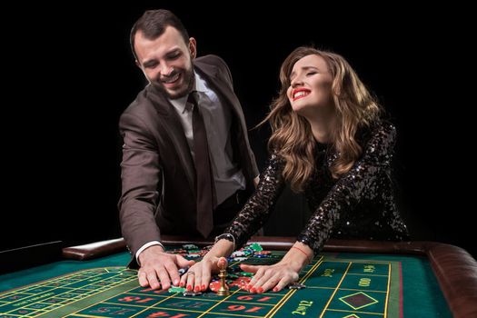 A man in a brown suit with a beautiful woman in a black dress is playing at a roulette table at a casino. Emotion of gambling players