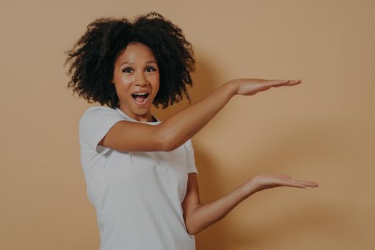 Excited Afro American beautiful young woman with black curly hair showing with hands example of size for advertising, isolated over dark beige background with copy space, positive emotions concept