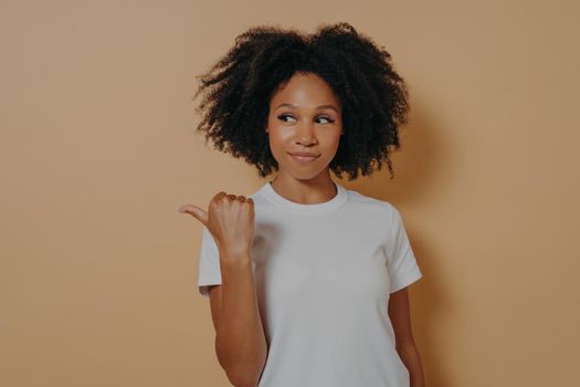 Pretty curly haired african woman points thumb aside, recommends promo and advertises product with glad expression, suggests visit this place while poses over beige background