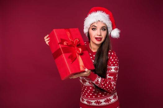 Young woman with long beautiful hair in Santa hat and New Year's dress holding Christmas present in her hands on red studio background. Christmas sale. Emotional brunette. Copy space