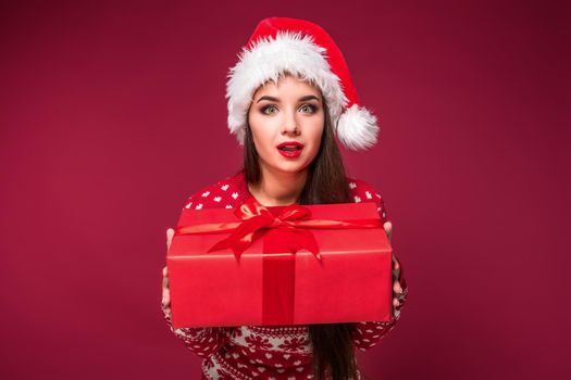 Young woman with long beautiful hair in Santa hat and New Year's dress holding Christmas present in her hands on red studio background. Christmas sale. Emotional brunette. Copy space