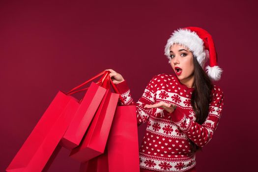 Young pretty woman or girl with long beautiful hair in Santa hat and New Year's dress holding red package on red studio background. Christmas sale. Emotional brunette. Copy space