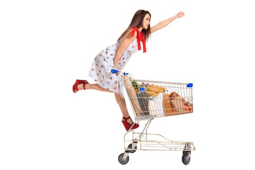 Woman with shopping cart full with products isolated over white background. A young woman rides for fun on an iron shopping basket, raising her hand up like a superman