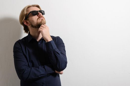 Thoughtful man in black sunglasses standing near a white wall