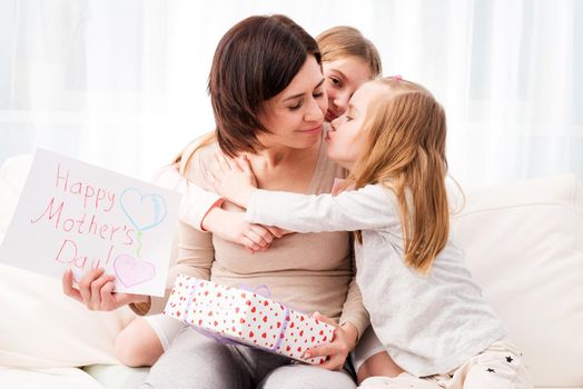 Two little sisters congratulate their mom with Happy mother's day. Children hugging and kissing mother and giving her creative hand-made drawing and gift box