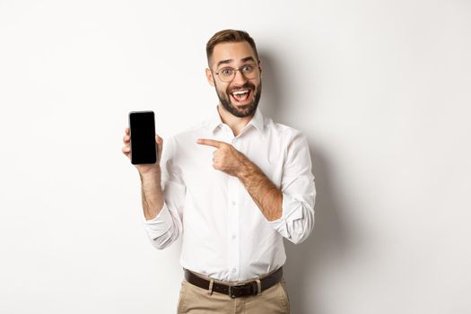 Excited handsome guy showing mobile phone, pointing finger at screen and smiling, standing against white background.