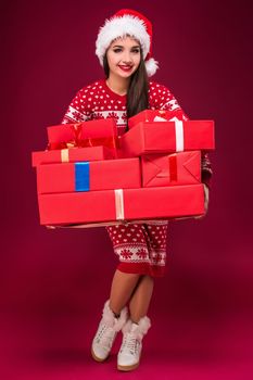 Amazed beautiful woman holding many presents on red background. Young woman in santa hat looks at camera. Christmas. Discounts. The shops. Sale