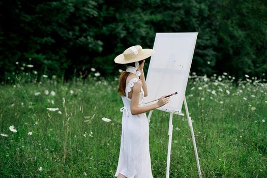 A woman in a white dress in a field with flowers paints a picture. High quality photo