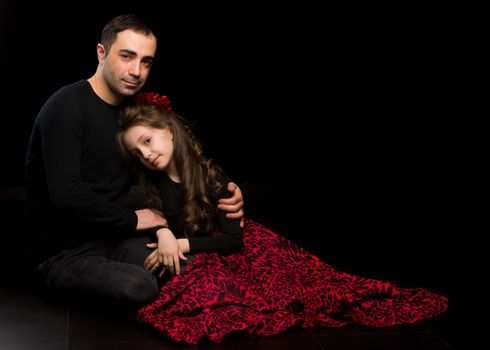 Portrait of Loving Father Hugging His Beautiful Daughter, Handsome Young Man Sitting on the Floor with his Adorable Girl, Happy Family Sitting Against Black Studio Background