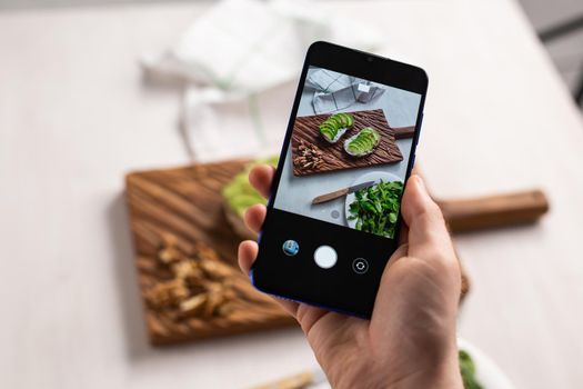 Hands take pictures on smartphone of two beautiful healthy sour cream and avocado sandwiches lying on board on the table. Social media and food