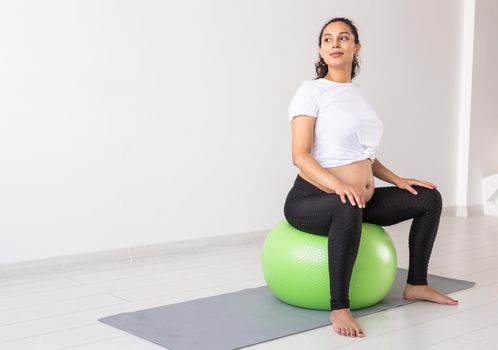 A young pregnant woman doing relaxation exercise using a fitness ball while sitting on a mat and holding her tummy