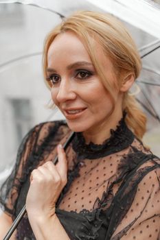 The blonde stands under a transparent umbrella during the rain. The fall season. Rear view. The woman is dressed in a black lace dress, her hair pulled back in a ponytail