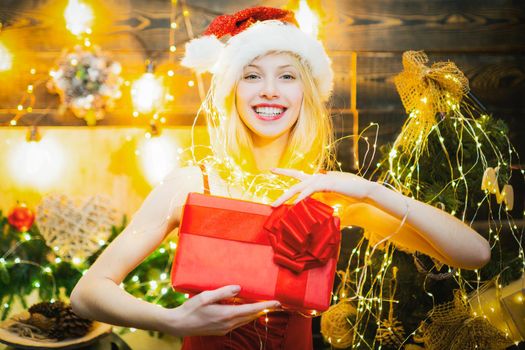 Beautiful young woman in Santa hat over christmas decorated interior background holding a gift or present. Winter shopping sales concept