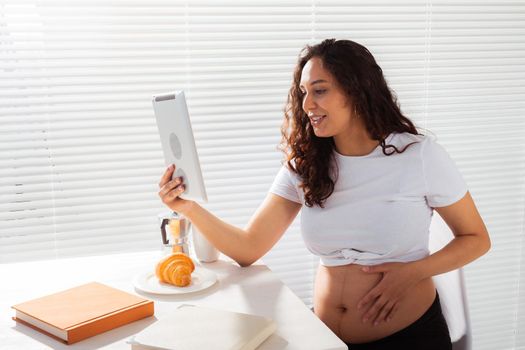 Happy pregnant young beautiful woman talking to mom using video call during morning breakfast. Communication and positive attitude during pregnancy