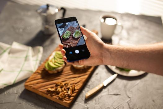 Hands take pictures on smartphone of two beautiful healthy sour cream and avocado sandwiches lying on board on the table. Social media and food