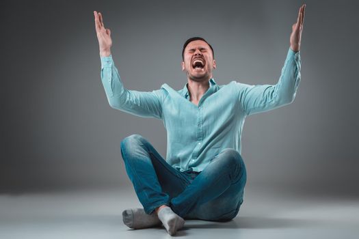 Handsome young man sitting on a floor with raised hands, isolated on gray background. Emotion concept