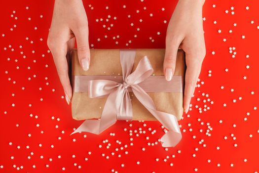 Nice female hands holding gift box wrapped in kraft paper with pink ribbon on festive red background with many snowflakes. Xmas and New Year postcard design. Giving love and warmth, Christmas concept