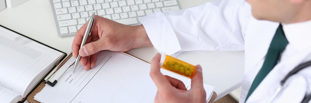 Male medicine doctor hand hold jar of pills and write prescription to patient at worktable. Panacea and life save, prescribing treatment, legal drug store concept. Empty form ready to be used