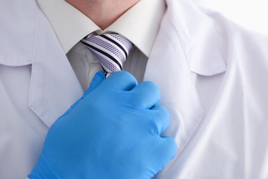 A male doctor in a dressing gown and shirt adjusts his tie with his hand in a blue glove