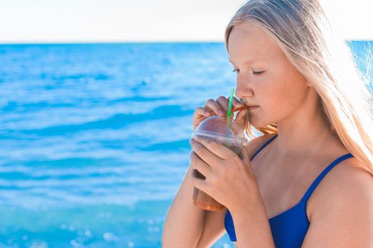 A young girl with blonde hair of European appearance, a teenager holds and drink a colored cold non-alcoholic cocktail in her hand against the background of the sea beach.