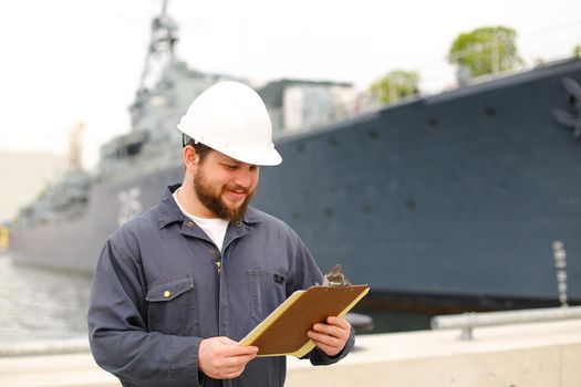 Deckhand wearing helmet and holding papers documents, standing on coast near vessel. Concept of maritime job and engineering department of marine team.