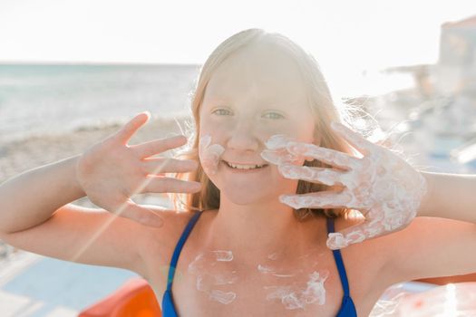 Portrait of a young positive teenage girl blonde of European appearance with sunscreen on her face and body against the background of a sea beach.