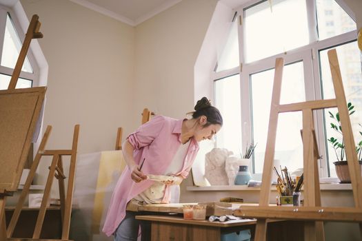 Cute woman paints on canvas in an art workshop. Artist creating picture. Art school or studio. Work with paints, brushes and easel. Hobby and leisure concept. Woman painter at workspace.