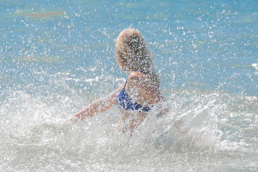 Young cheerful girl teenager blonde makes splashes of water with her hands in the sea.