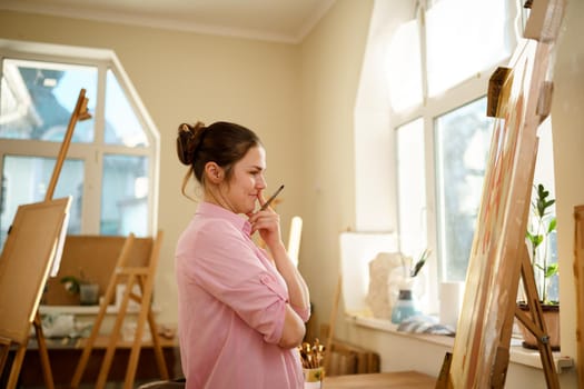 Cute woman paints on canvas in an art workshop. Artist creating picture. Art school or studio. Work with paints, brushes and easel. Hobby and leisure concept. Woman painter at workspace.